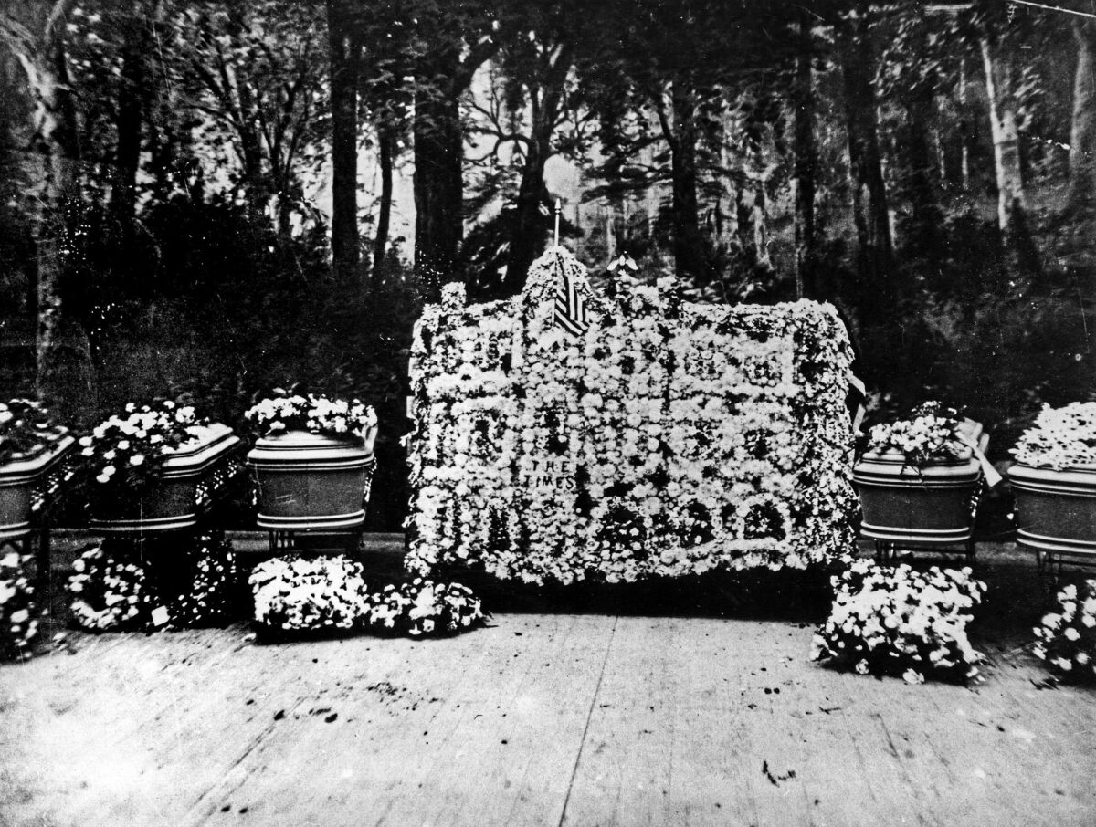 Caskets at funeral services for the dead in the bombing of the Los Angeles Times building. The huge floral display is a reproduction of the Los Angeles Times building. The background is a painted mural of a forest scene.