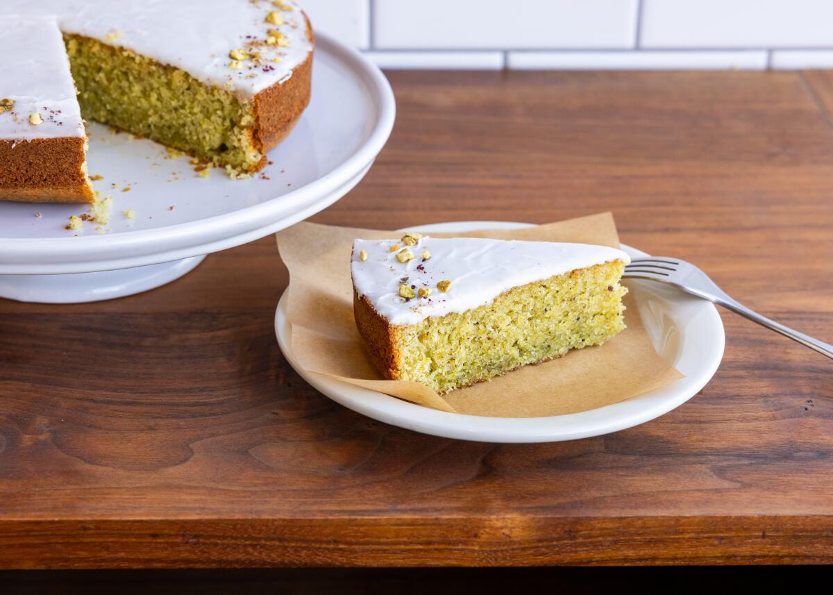 A triangular slice of pistachio olive oil cake with lemon glaze on a small plate next to the remaining cake on a cake stand