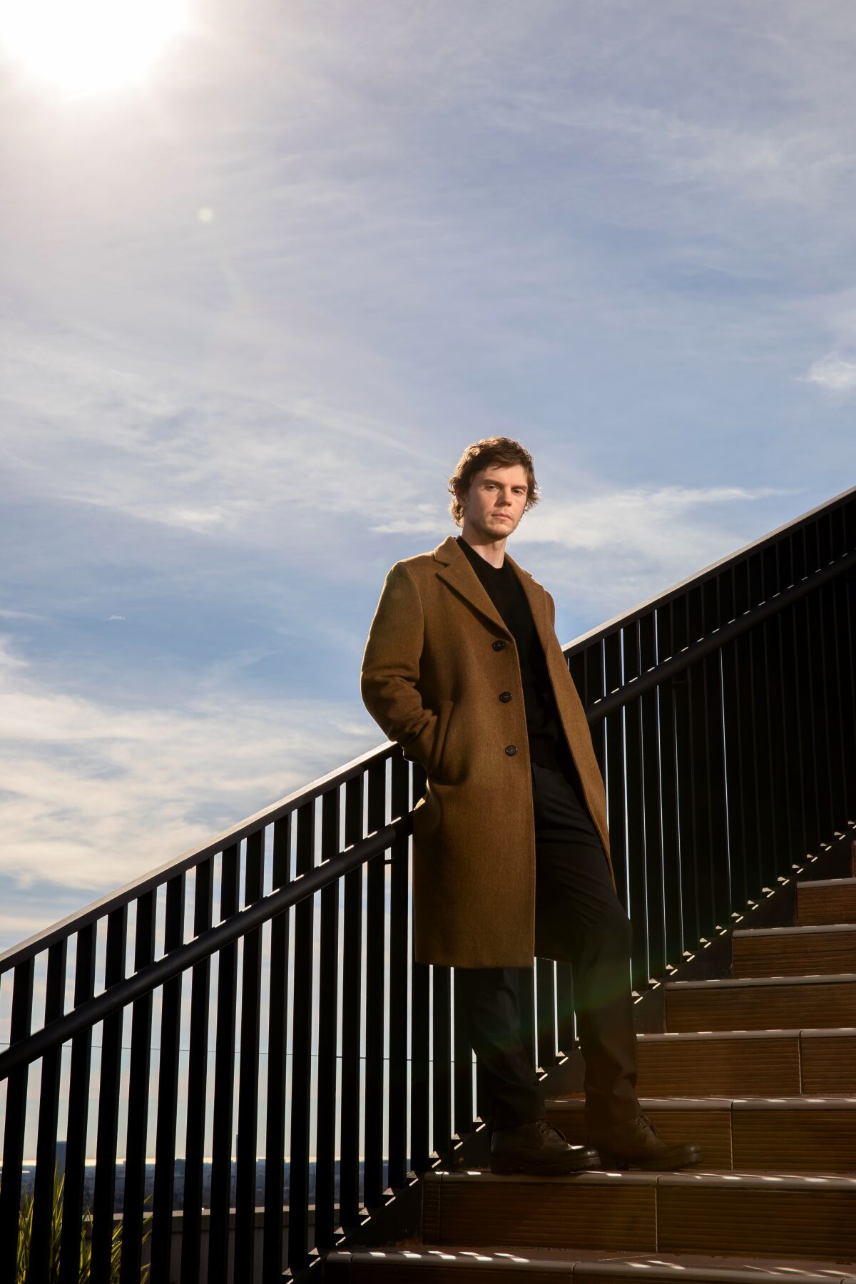 Evan Peters leans against a stairway railing against a cloudy blue sky.