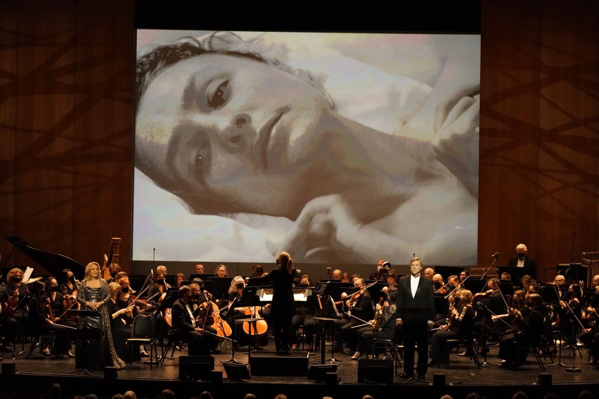 A projection of a woman, her head on a pillow, and a live orchestra and singers in front of the screen.
