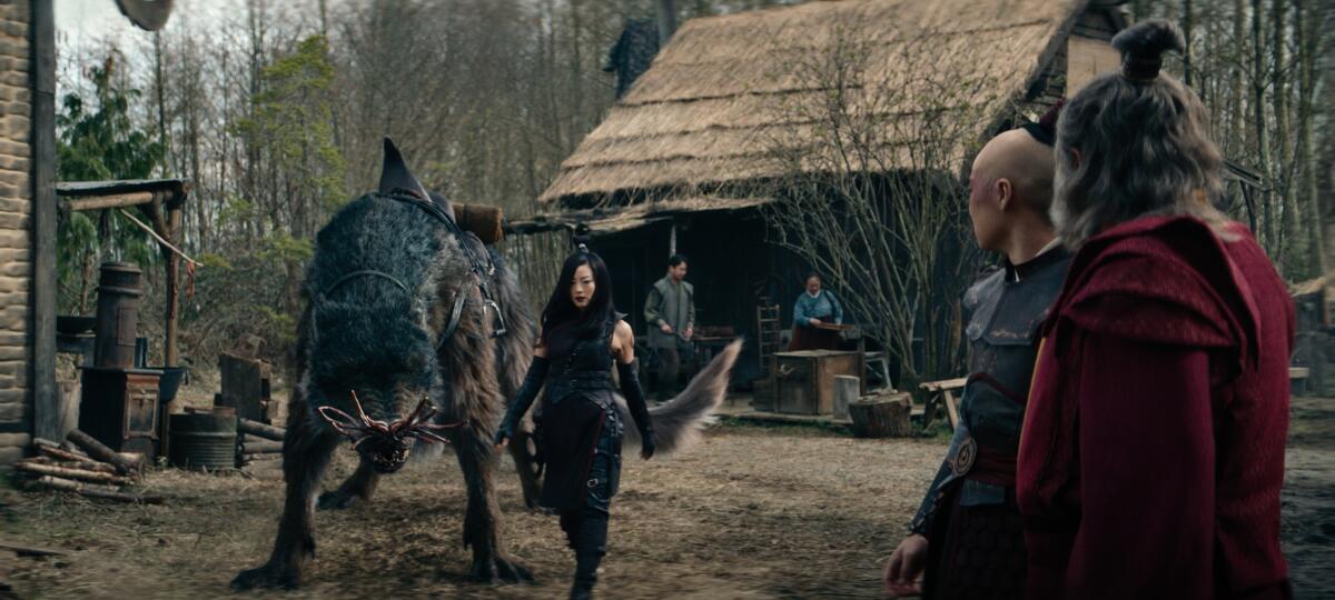 A woman leads a winged creature into a village in the forest while two men look on.
