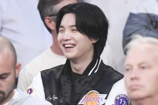 A young Korean man with dark medium-length hair sitting and smiling wearing a black letterman jacket with the Lakers logo