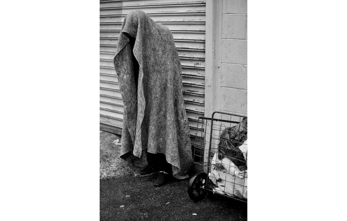 A person stands beneath a blanket in the rain trying to keep dry, on San Pedro Street on skid row.