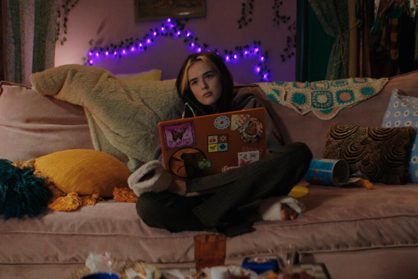 A young woman with a laptop sits on a couch in the movie "Not Okay."