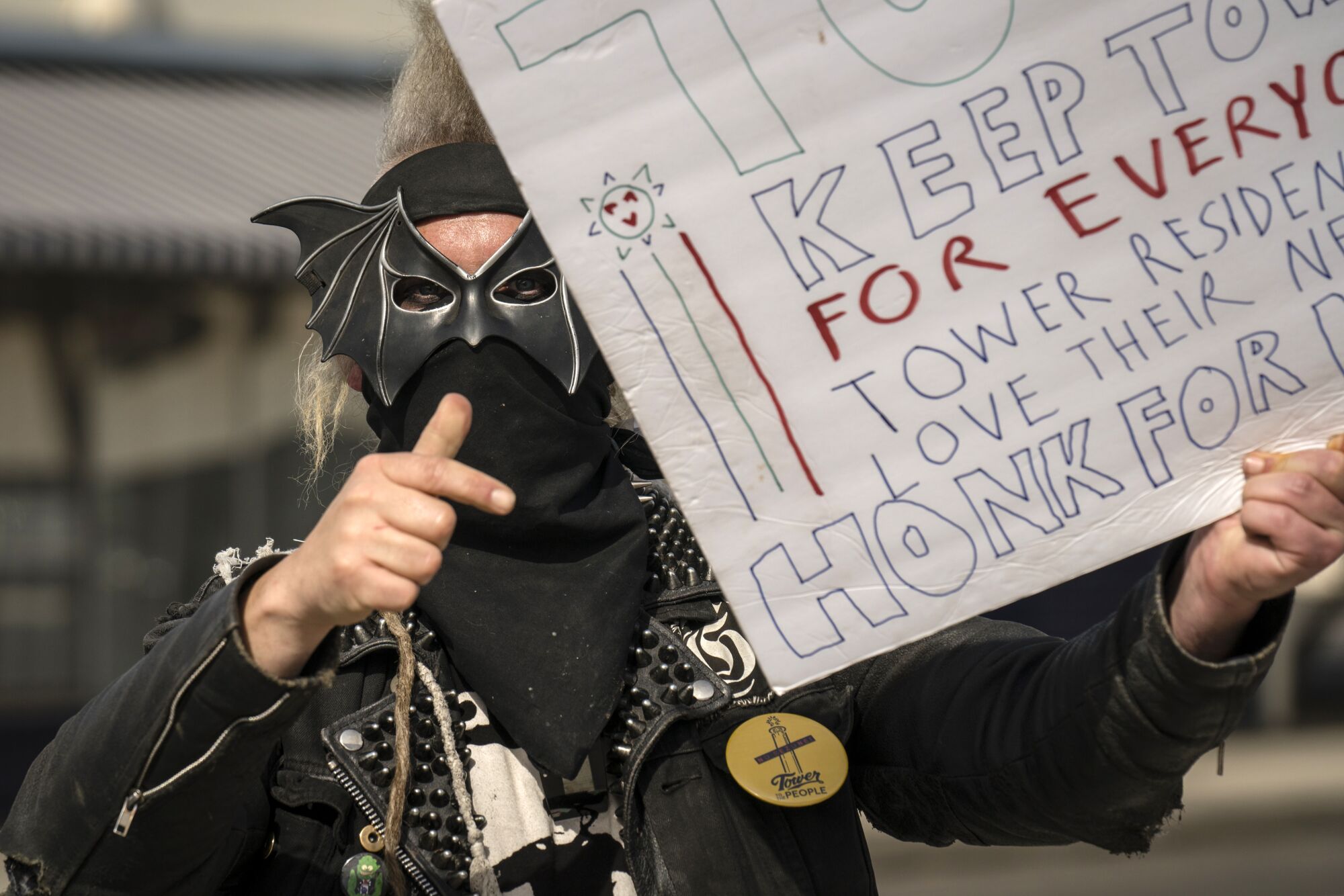 A man in a leather jacket and a bat mask holds a hand-written sign