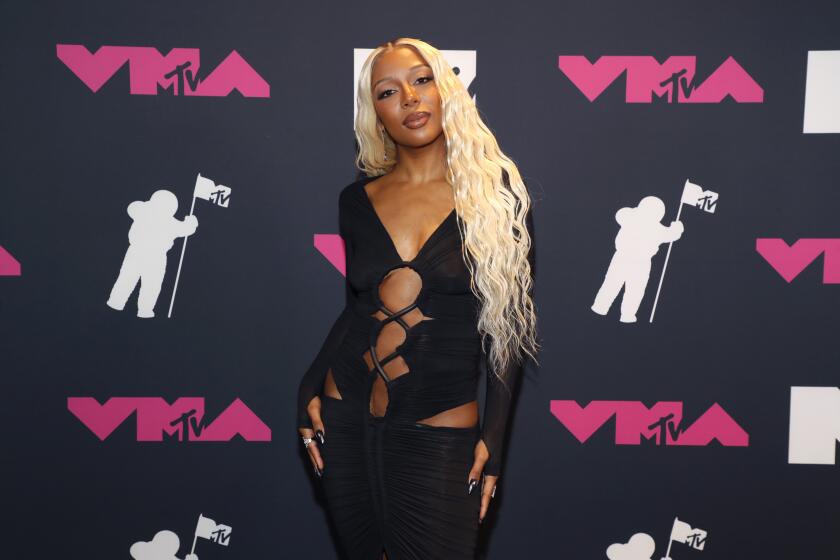 A black woman with wavy blond hair in a revealing, tight-fitting black dress with cut-out details 