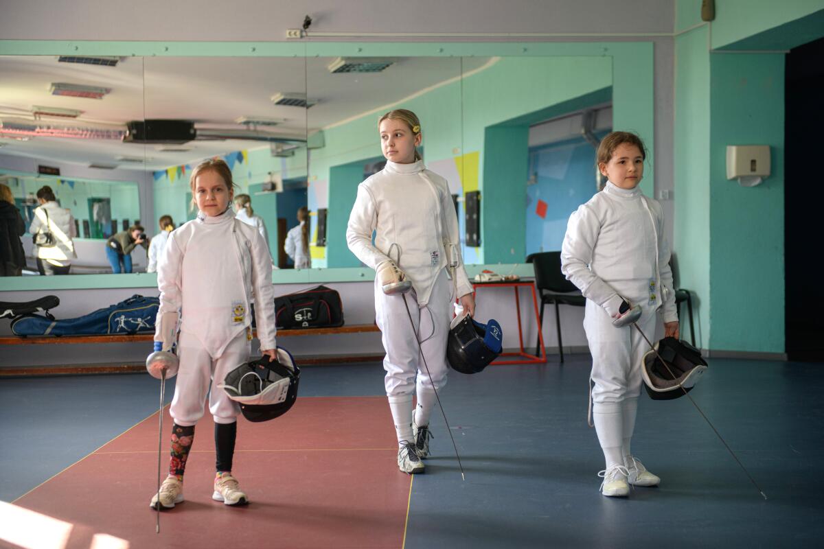 Three girls stand in fencing uniforms, holding helmets and equipment.
