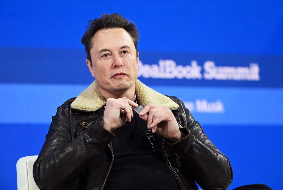Elon Musk sits before a backdrop that says "DealBook Summit"