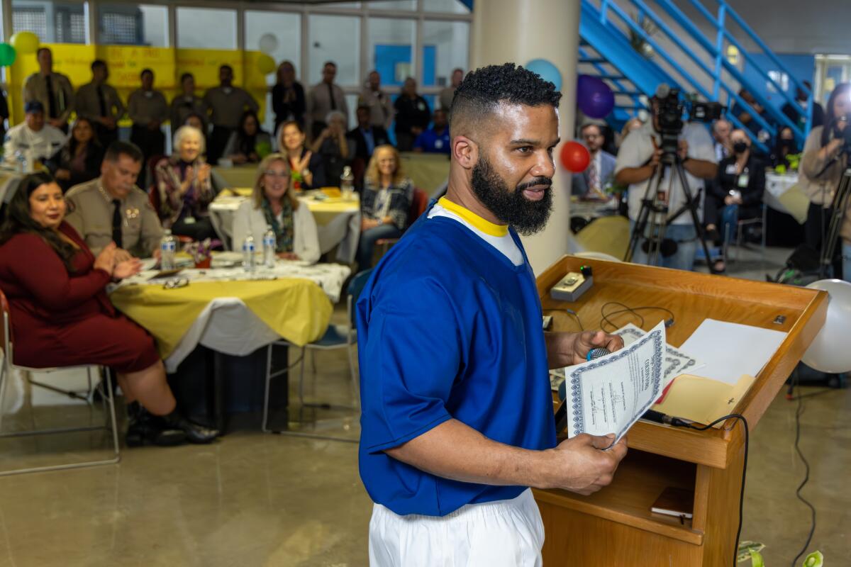 A bearded man with dark hair, in a blue shirt, holds a certificate at a lectern, surrounded by people seated at tables