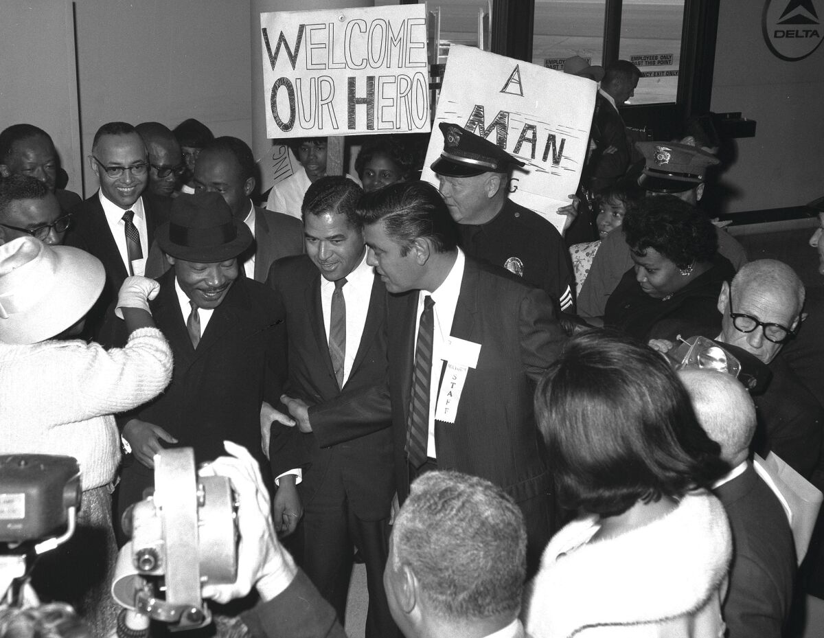 Martin Luther King Jr. is surrounded by people, some with signs. One sign says "Welcome Our Hero."