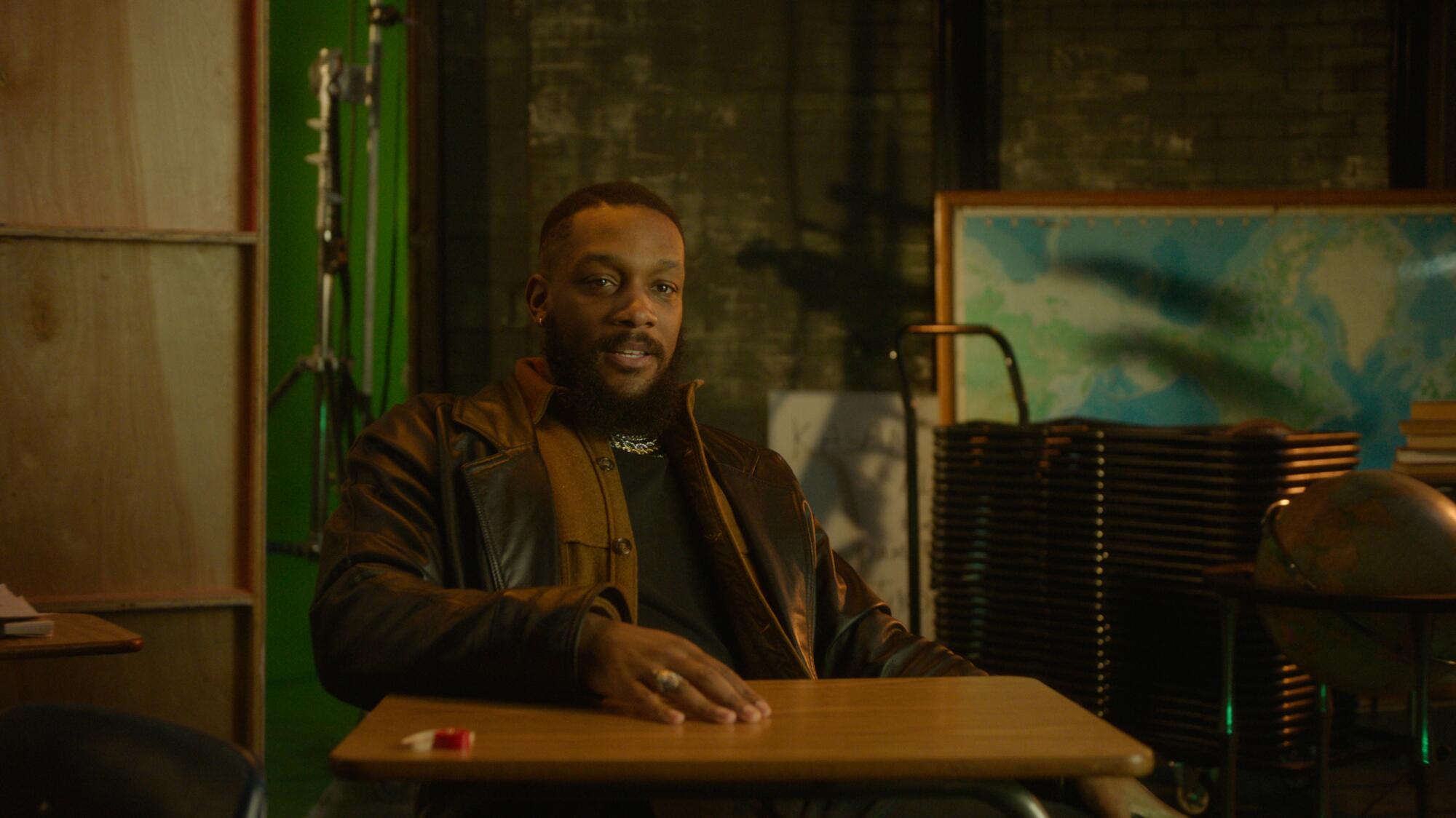 Bryan Hearne, in a leather jacket, sits at a desk on a TV show set.