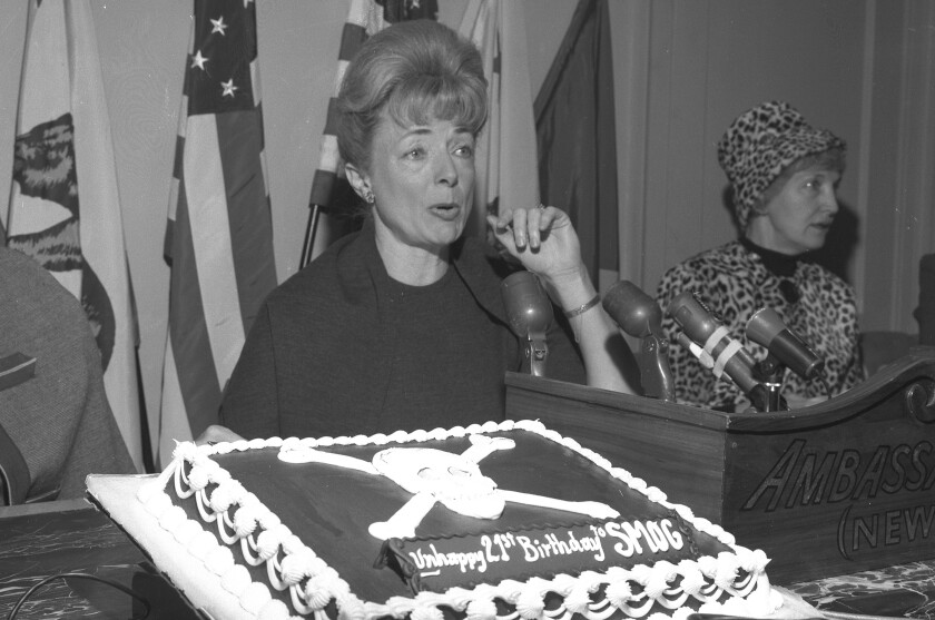 Two women sit at a table in front of microphones and a cake with a skull and crossbones.