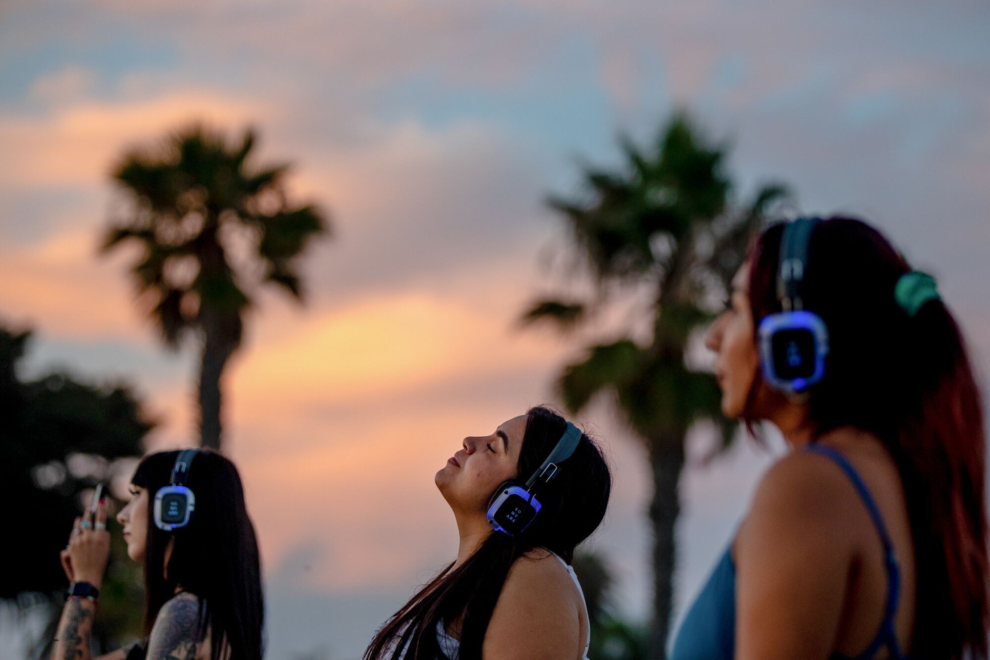 Side view of three people standing with headphones on, eyes closed, against a sunset and palm tree background.