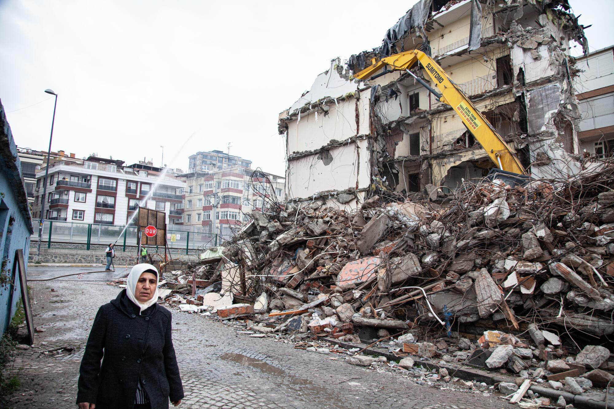 A woman in dark clothes and white headscarf walks near a building under demolition, with rubble piled high 