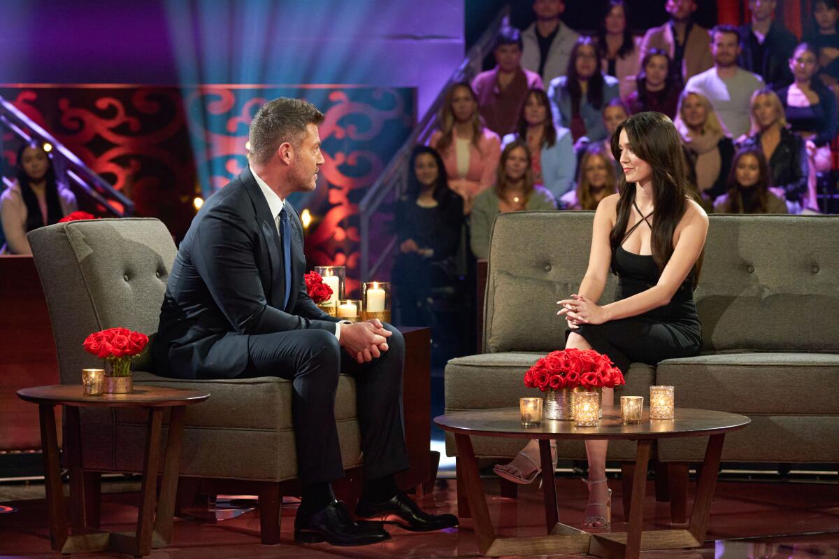 A man and a woman in eveningwear sitting on couches on a TV set.