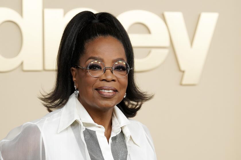Oprah Winfrey with her hair in a high ponytail, glasses and a sheer white blouse smiling against a gold background