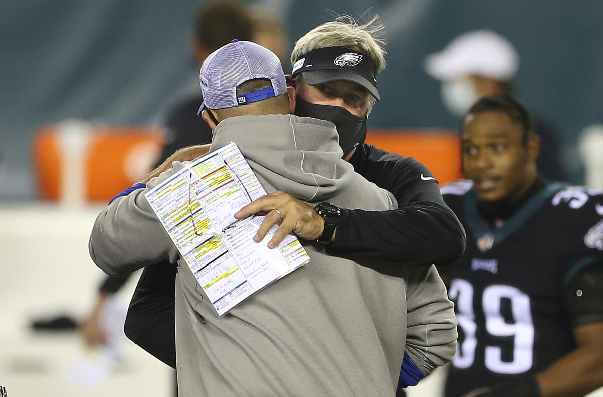 On the field, a masked Doug Pederson, game notes in his hand, hugs Joe Judge.