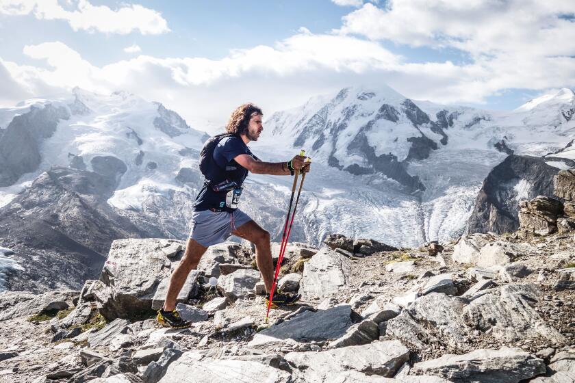 A man in T-shirt and shorts holds walking sticks as he travels along a rocky path, with snow-covered mountains behind him. 