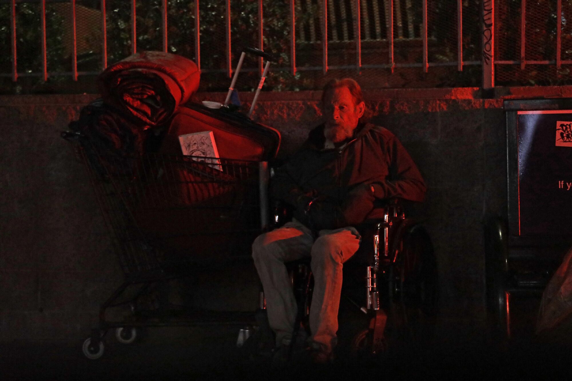 An elderly homeless man sits in his wheelchair next to his belongings on the sidewalk at night.