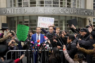 A bearded man in suit and tie speaks before microphones while standing behind barricades, surrounded by people