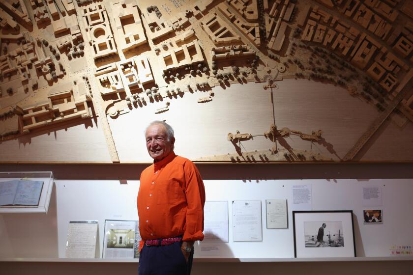 Richard Rogers, wearing a bright orange shirt, stands before a wooden model of a city suspended against a wall