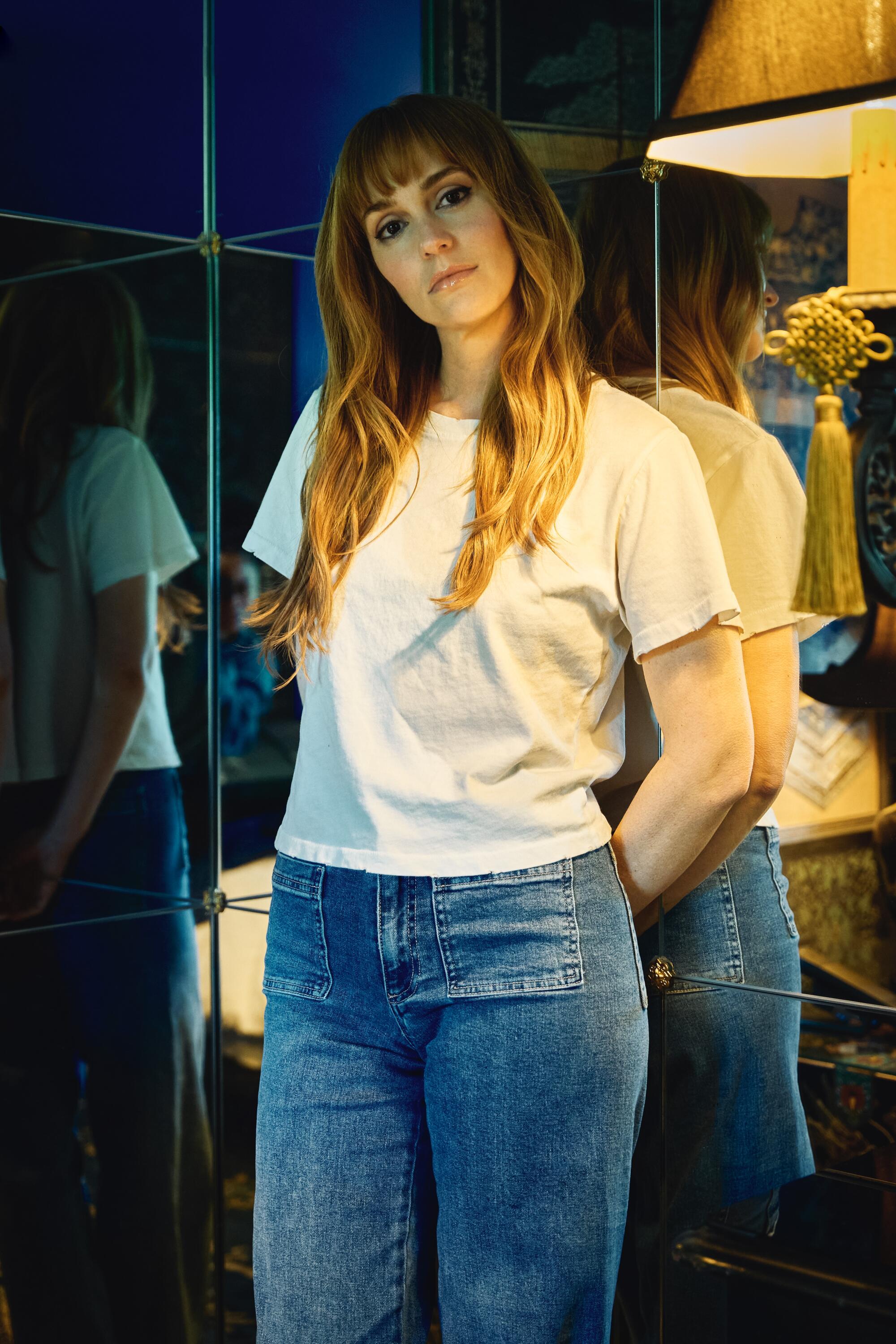 A pistillate successful achromatic T-shirt and bluish jeans stands against a mirror, hands down her, caput tilted