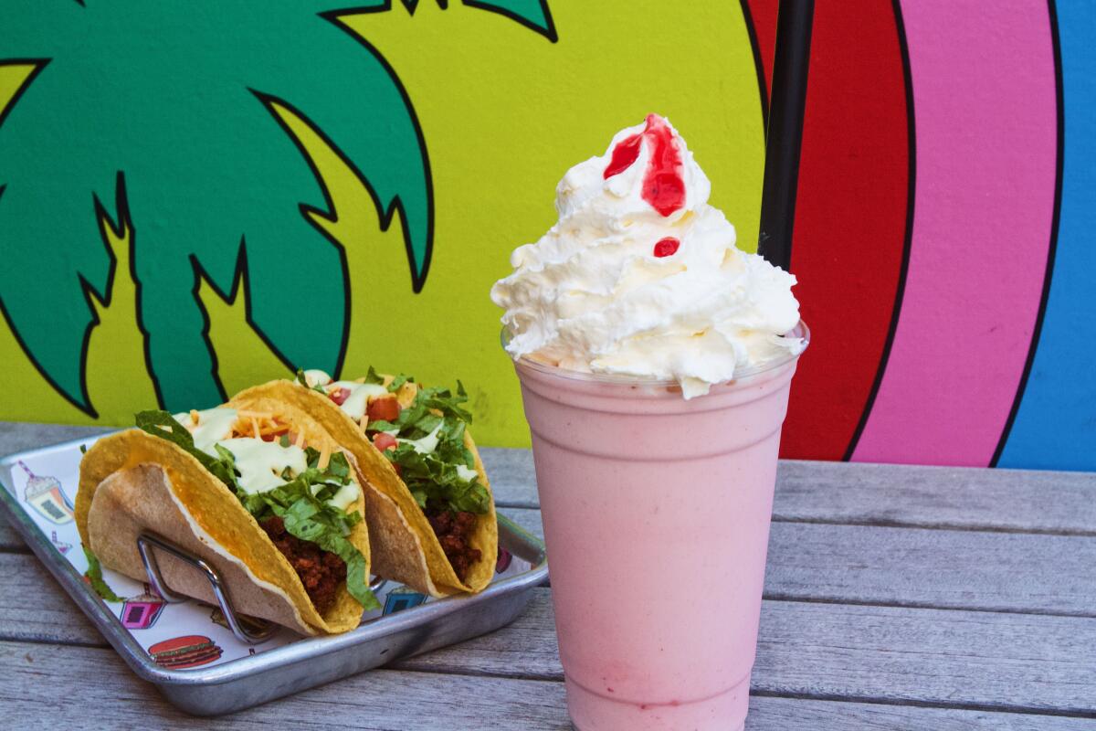 A strawberry shake and two tacos from Nomoo sit against a palm tree mural. Strawberry syrup trickles over coconut whip.