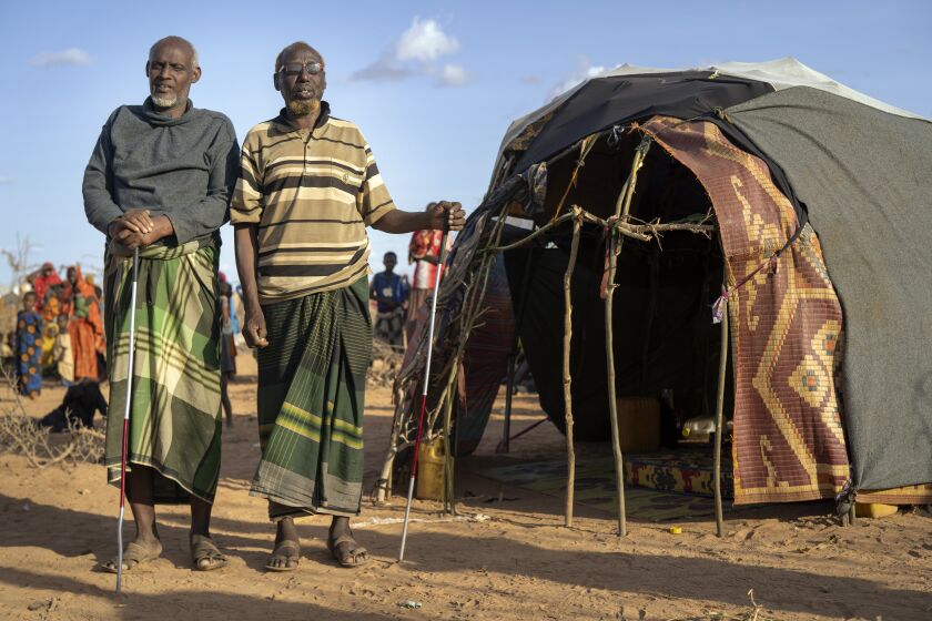 Mohamed Kheir Issack, 80, right, and and Issack Farow Hassan, 75, stand outside Issack's shelter at a camp for displaced people on the outskirts of Dollow, Somalia on Tuesday, Sept. 20, 2022. The two blind men are friends and as close as brothers, gripping each other's hands in their mutual darkness as tightly as they hold their canes. Near the end of their lives, the most alarming drought in more than half a century in Somalia has stripped them of their animals and homes. (AP Photo/Jerome Delay)