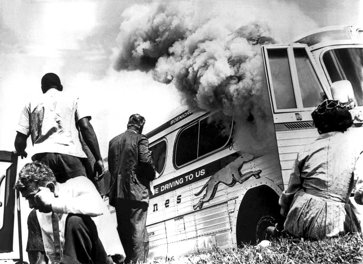 People sit and stand outside a bus that is on fire, with smoke coming out the door.