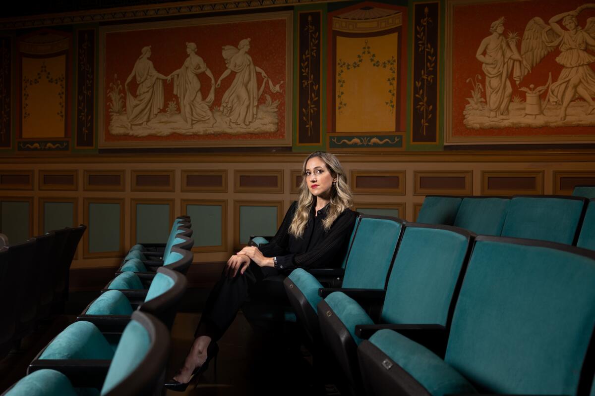 Melissa Barak, a white woman in a dark suit, sits in a row of velvety theater seats before murals of Greek muses. 