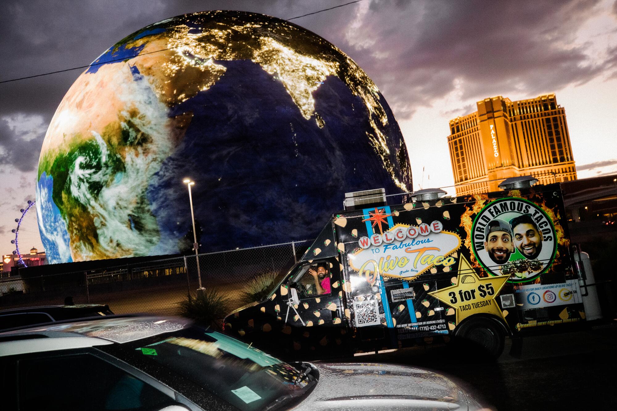 A taco truck is parked in front of Sphere at dusk, with the building featuring projections of Earth.