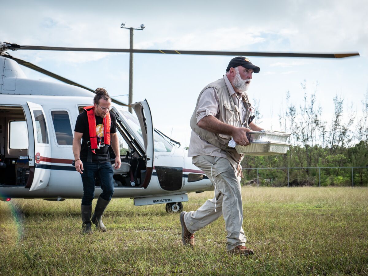 Two men, one carrying trays of food, exit a helicopter in a grassy field.