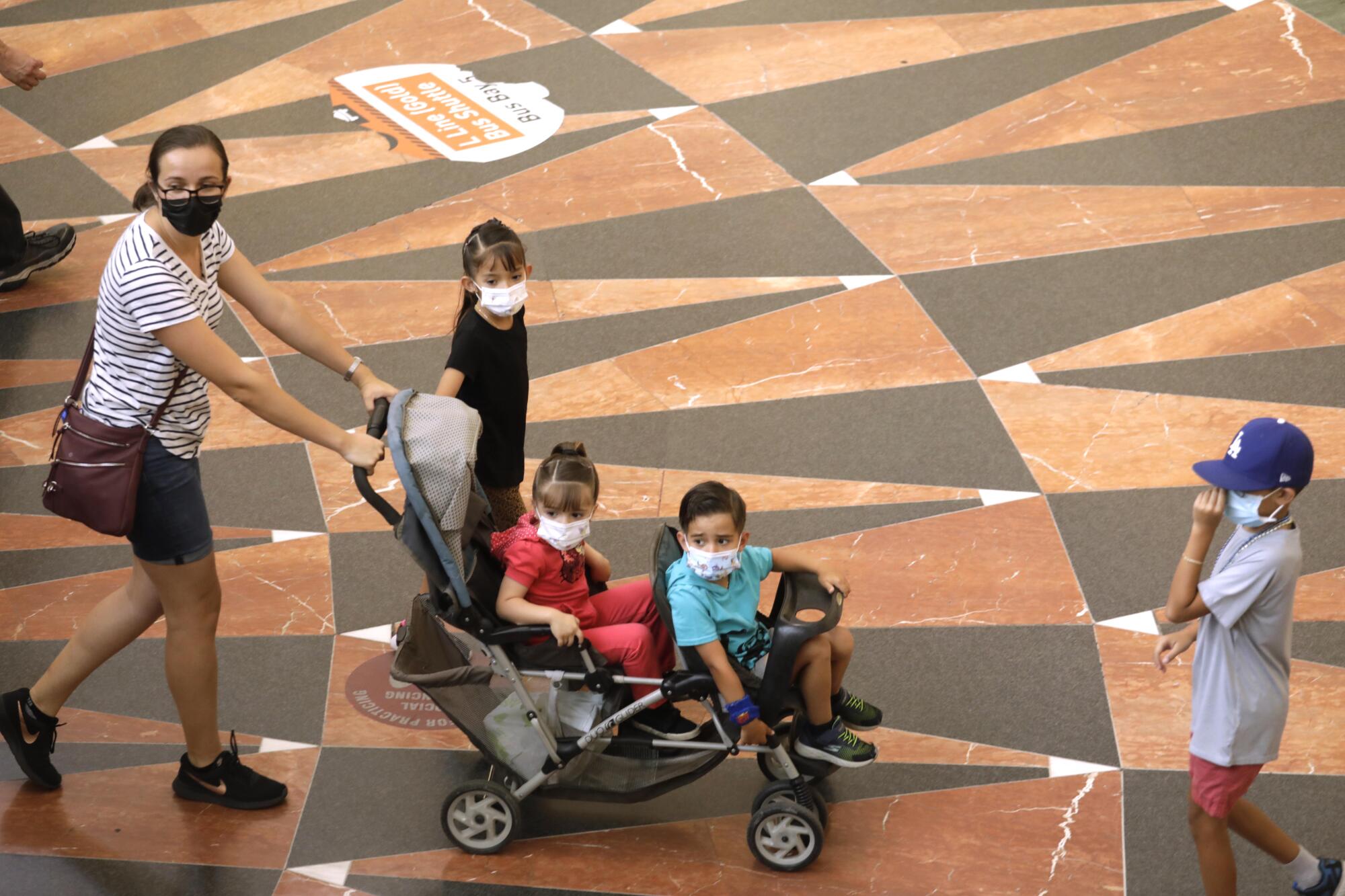 A woman pushes a double stroller with children. All wear masks.