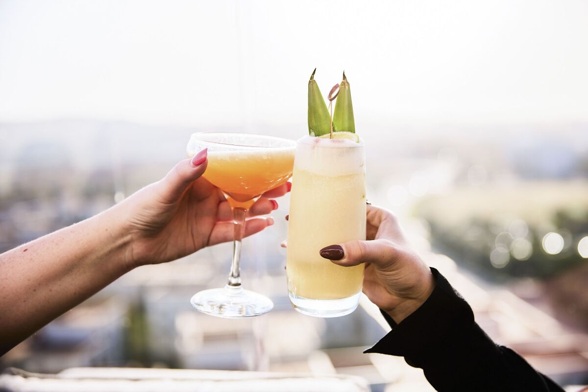 two hands clink cocktail glasses at an outdoor rooftop bar