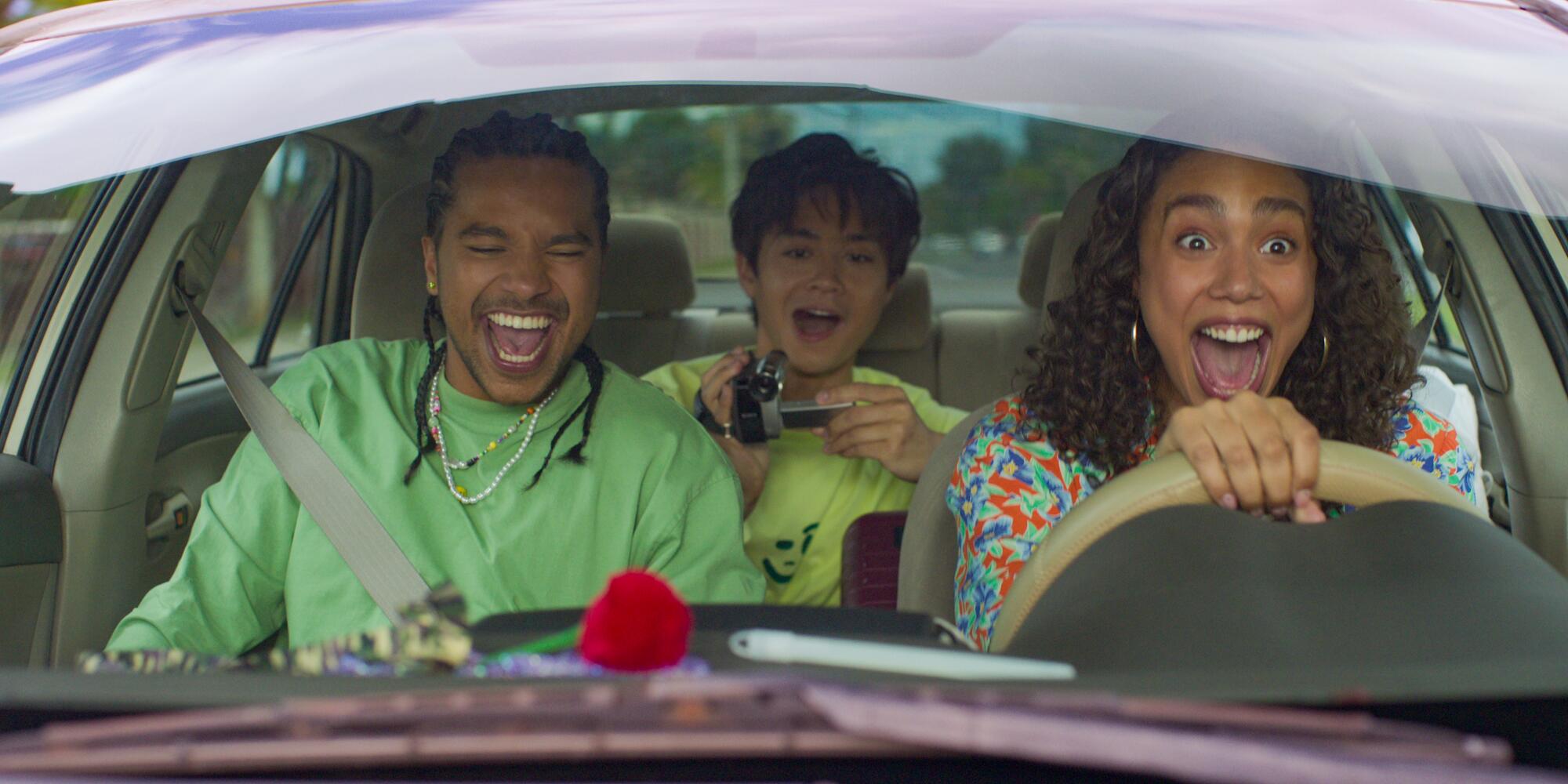 A man and a woman with wide smiles ride in the front seat of a car while another man holds a video camera in the backset.