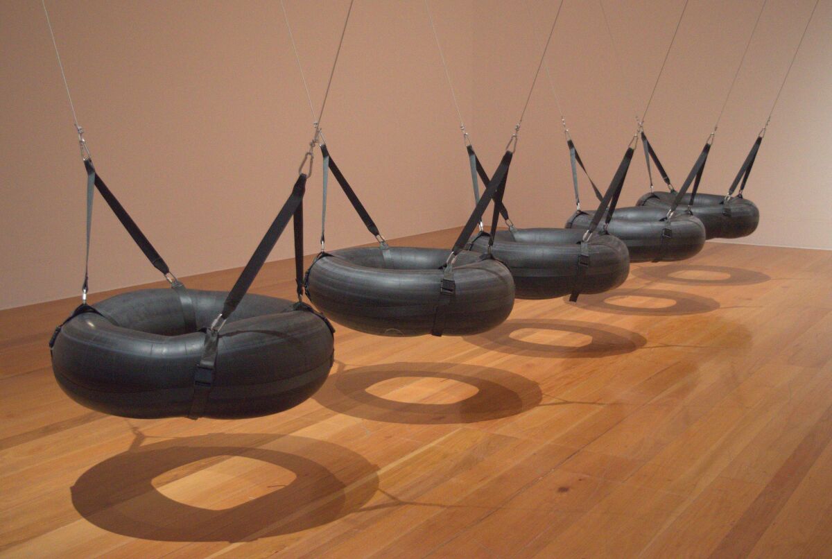 An installation in a gallery shows a row of black inner tubes suspended like swings from the ceiling