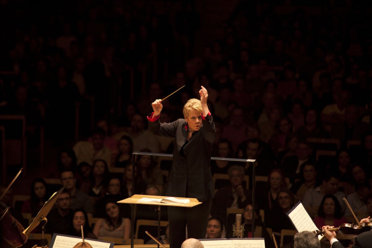 A woman wearing a dark suit holds up a baton and gestures with her other arm while conducting an orchestra