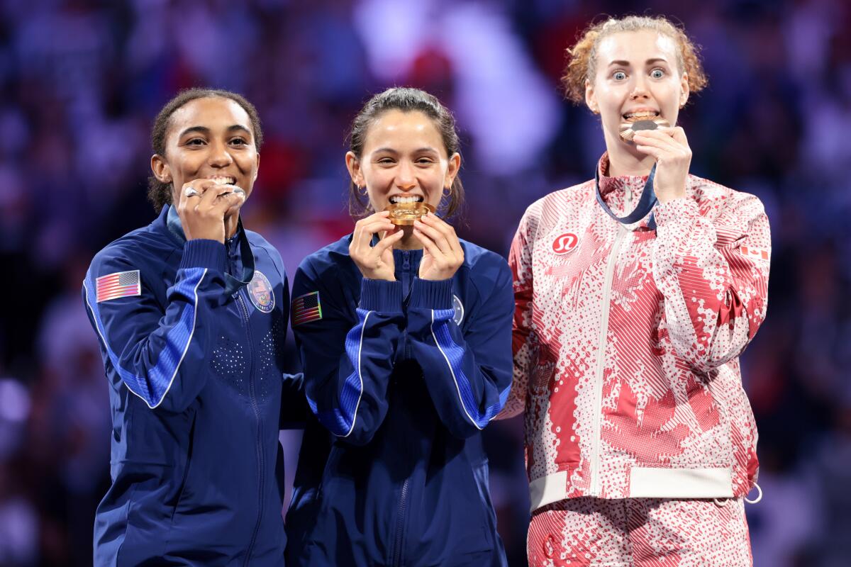 Lee Kiefer, center, of the U.S. bites her gold medal next to Lauren Scruggs and Eleanor Harvey.