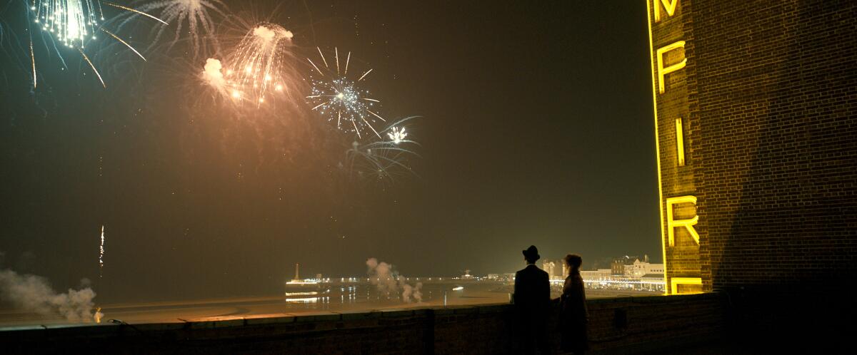 Two silhouetted figures watch fireworks from a rooftop beside a lighted movie marquee.