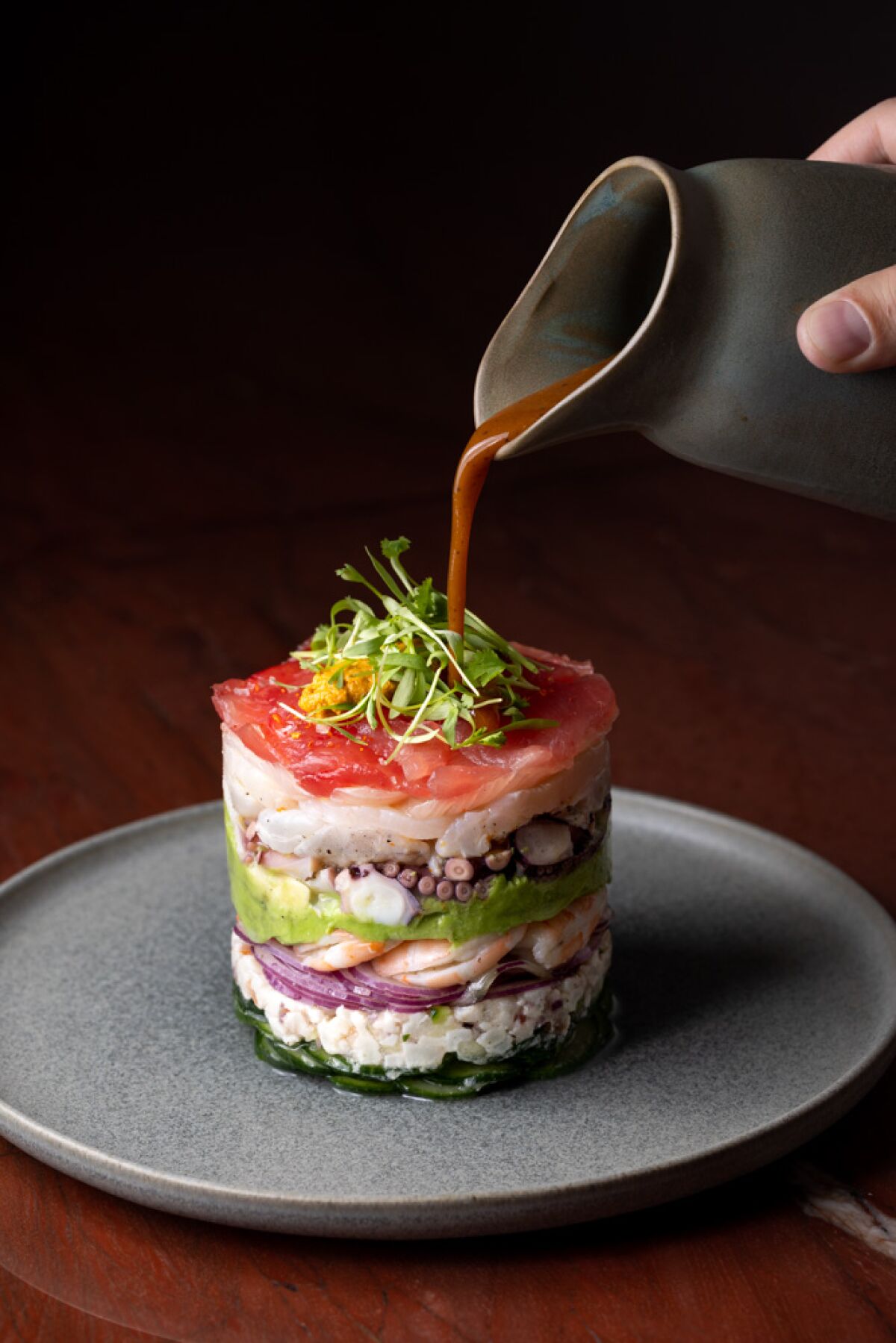 A hand pours sauce over a tower of stacked shrimp, tuna, octopus, scallop and uni 