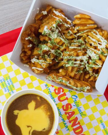 An overhead photo of a box of Katsu Sando waffle fries topped with furikake and mayo. A side of curry with cheese sauce.