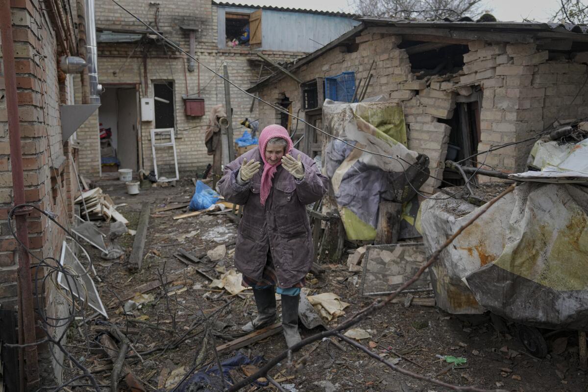 A woman with a pink head scarf, in winter clothes, boots and gloves, stands in the midst of damaged brick buildings 
