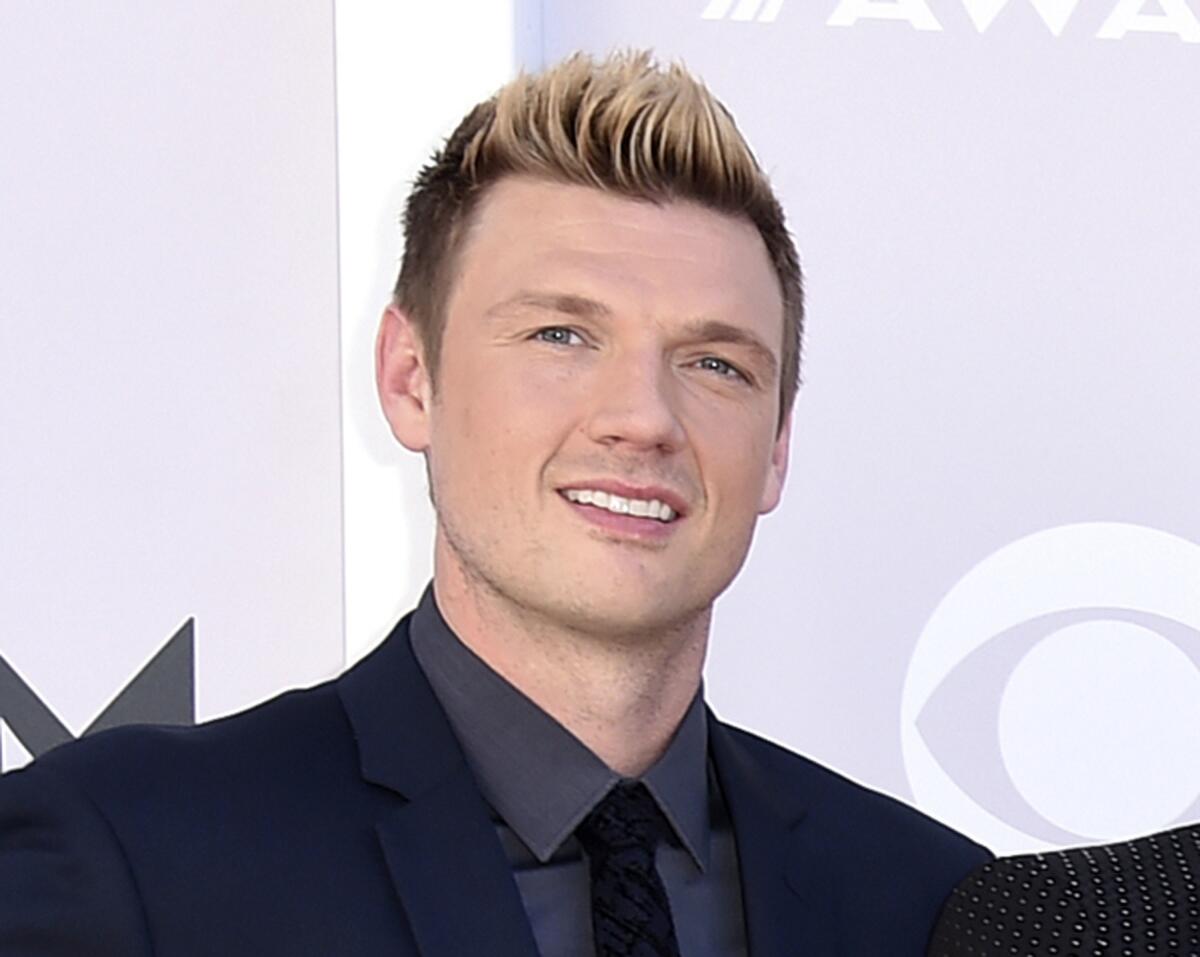 Nick Carter wears a black suit jacket with a dark shirt and tie in front of a backdrop