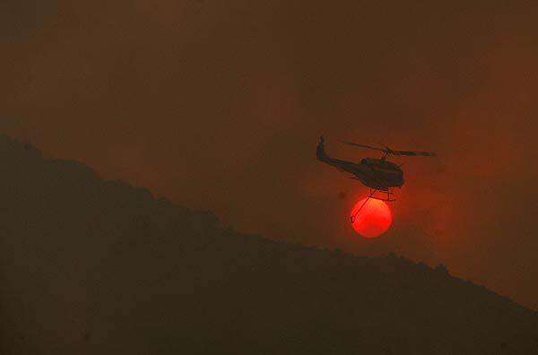 A helicopter finishes a water drop and flies over the setting sun over the town of Acton.