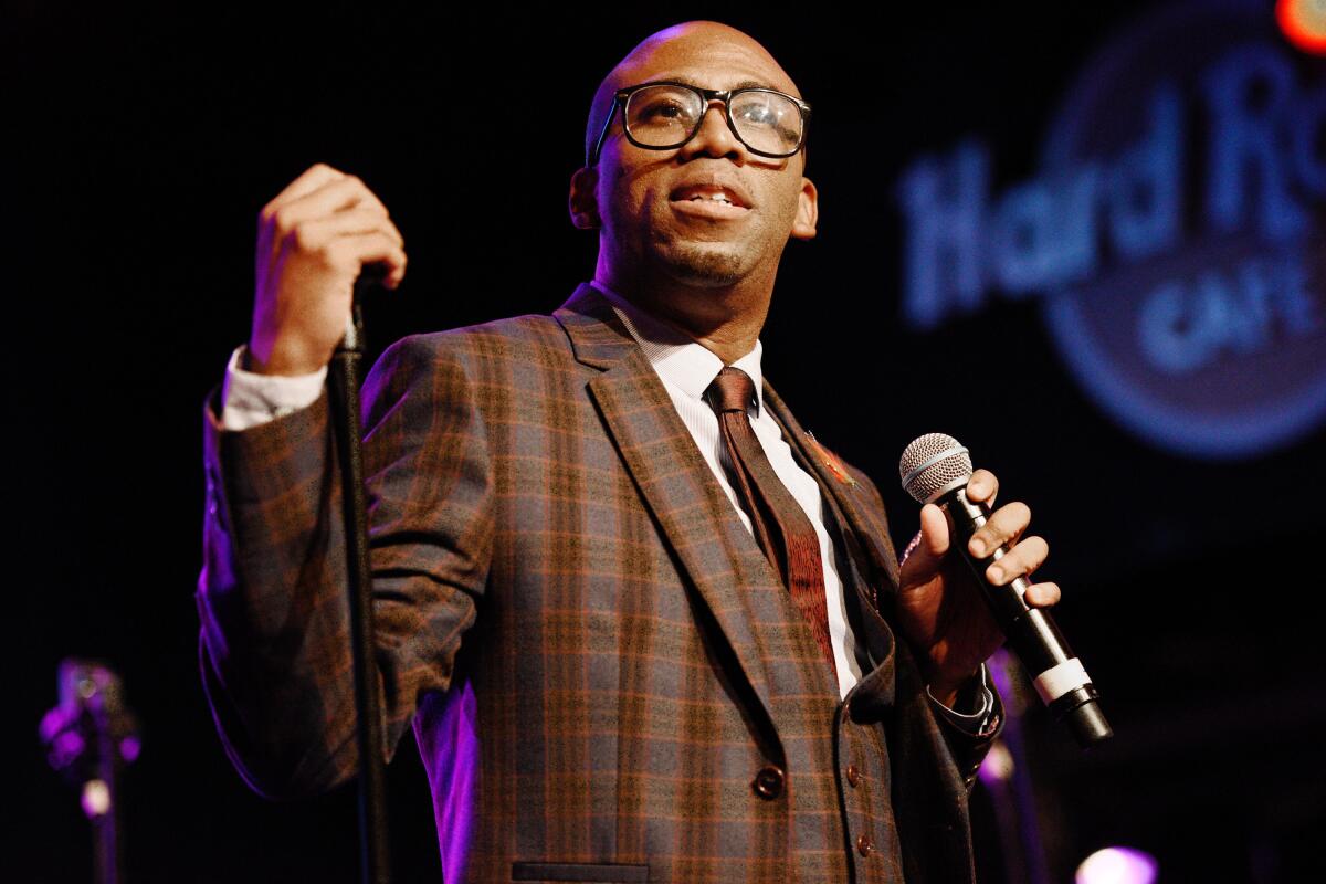 A bald man wearing glasses and a plaid suit holding a microphone on a stage