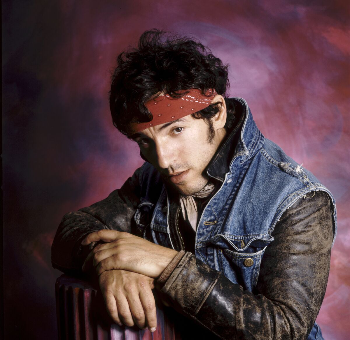 Bruce Springsteen, circa 1980, wearing a denim vest over a leather jacket with a red bandanna wrapped around his head.