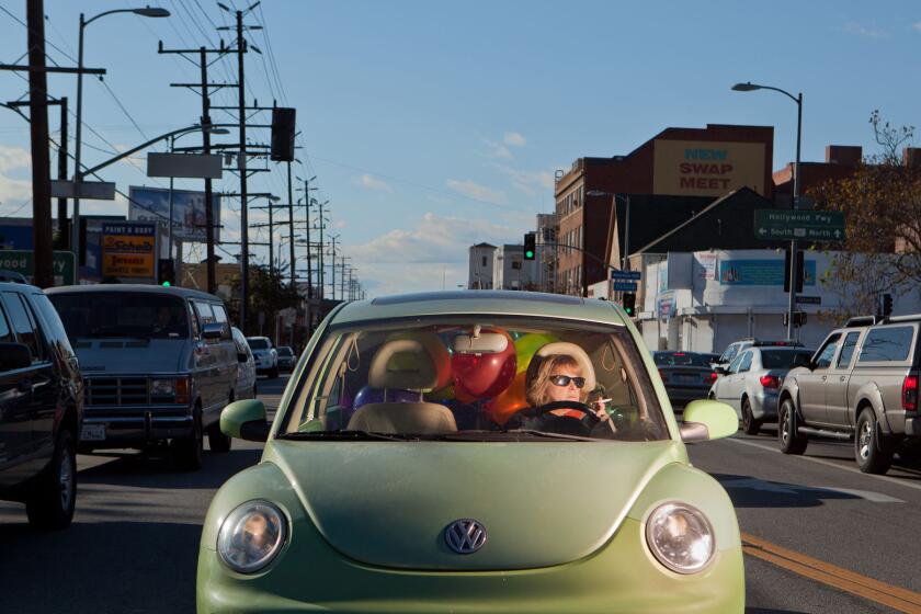 A woman wearing sunglasses and holding a cigarette sits at the wheel of a green VW Beetle with a backseat full of balloons.