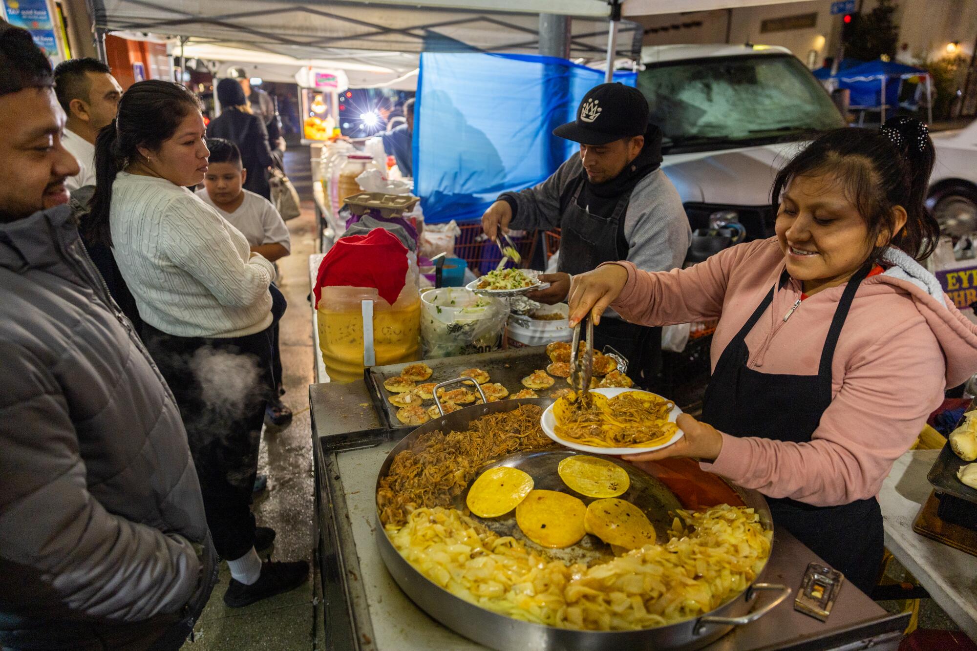 Customers wait for their food as a woman serves tacos at an outdoor market