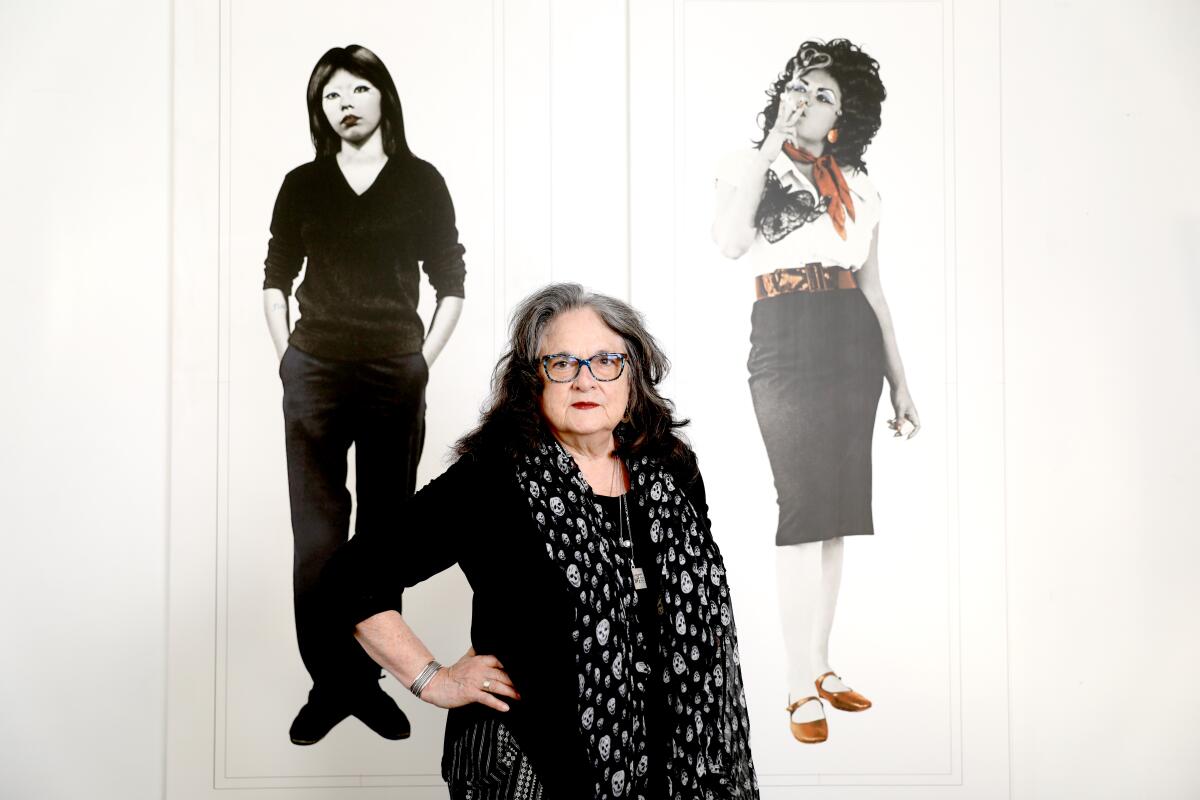 Artist Judy Baca stands before two images of women on a wall.