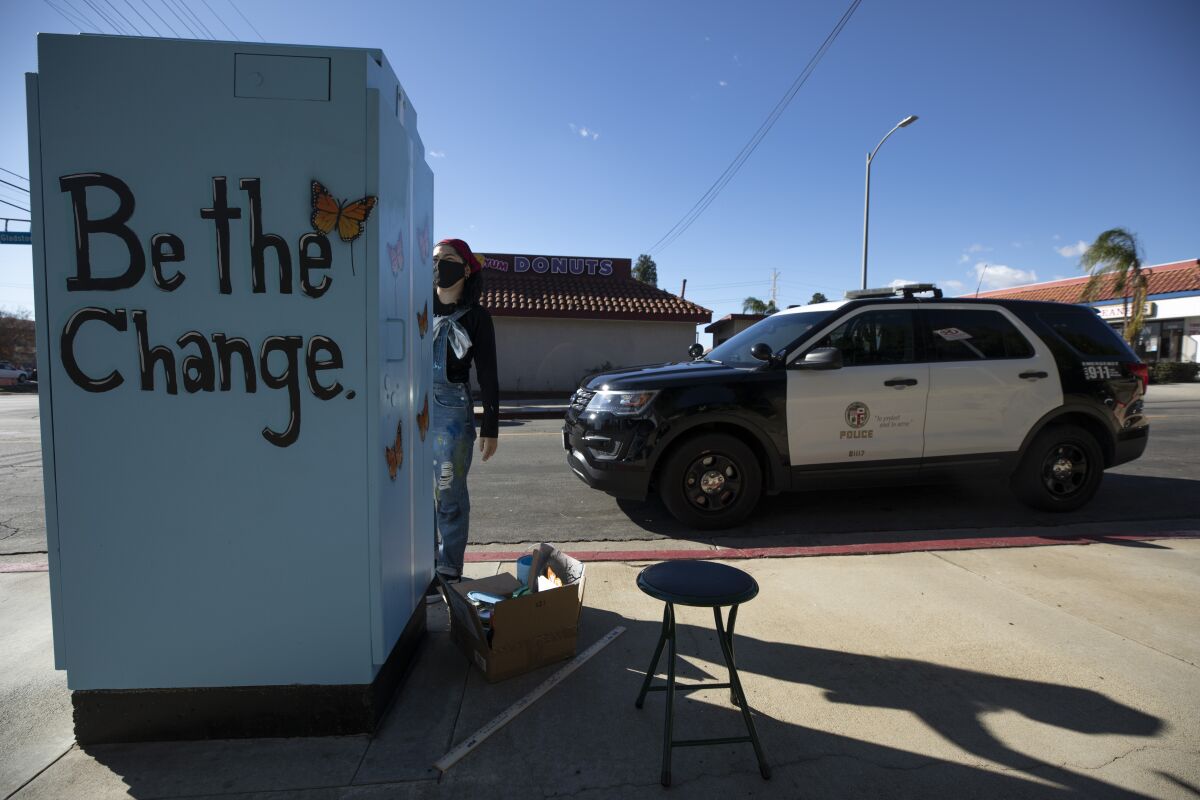 A police vehicle is parked at the curb alongside a young artist working on a mural.